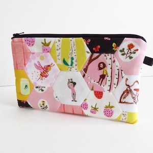 May include: A quilted pouch with a black zipper and pull tab. The pouch is made of colorful, patterned fabric with hexagonal patches. The patterns include bees, strawberries, and a girl under a tree. The pouch is approximately 9 inches wide and 6 inches tall.
