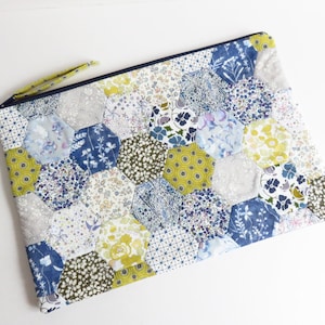 May include: A quilted clutch featuring a patchwork design of hexagonal fabric pieces in various floral patterns. The color palette includes shades of blue, yellow, and white. A navy blue zipper and a small fabric loop complete the design.