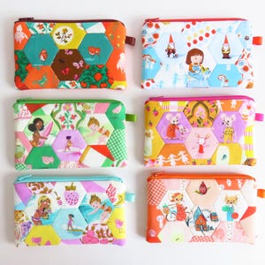 May include: Six colorful, patterned zipper pouches with hexagon patchwork designs. Each pouch features unique illustrations of gnomes, girls, and animals. The pouches have a rectangular shape with a zipper closure and a small loop on the side.