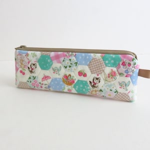 May include: A rectangular pencil case with a zip closure. The case features a patchwork design of hexagonal fabric shapes in various colours and patterns, including floral, gingham, and cat motifs. A small, tan fabric loop is attached to the side.