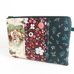 May include: A green zippered pouch with a patchwork design. The front features three panels of fabric: a white panel with a vintage illustration of a house in the woods, a burgundy panel with a floral pattern, and a dark green panel with a small floral pattern.