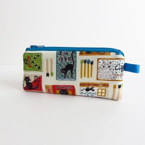 Puede incluir: Un estuche rectangular con cremallera y tirador azules. El estuche está estampado con ilustraciones de estilo japonés de gatos negros, cerillas y texto sobre un fondo crema. La cremallera es azul.