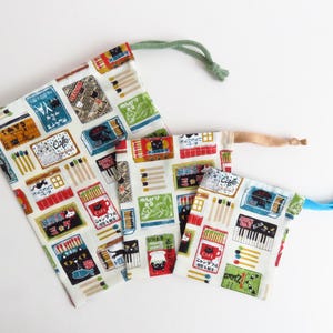 Puede incluir: Tres bolsas con cordón con fondo blanco y un patrón repetido de cajas de cerillas, gatos y letreros de café en rojo, verde, azul y marrón. Las bolsas tienen cordones verdes, de madera y azules.