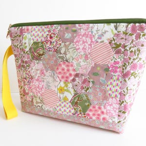 May include: A quilted pouch with a patchwork design featuring pink floral and geometric patterns. The pouch has a green zipper and a yellow handle. The bag is made of fabric and is likely used for carrying items.