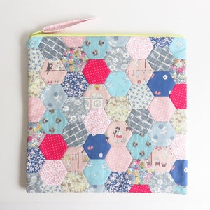 May include: A patchwork fabric zippered pouch with a hexagonal pattern in shades of pink, blue, and red. The pouch is made of various fabrics with different patterns, including floral, geometric, and animal prints.