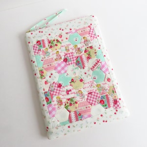 May include: A rectangular quilted pouch with a handle, featuring a patchwork design of hexagonal shapes. The fabric incorporates pink, mint green, and white patterns with strawberries, cherries, and illustrations of children. The edges are trimmed with a floral print.