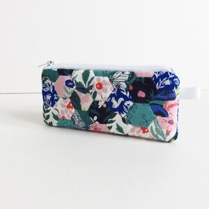 May include: A small zippered pouch with a white zipper and a white pull tab. The pouch is made of fabric with a patchwork design of hexagons in shades of blue, green, pink, and white. The fabric features floral and butterfly patterns.