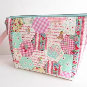 Puede incluir: Un estuche con cremallera de patchwork con un asa rosa claro. La bolsa presenta una variedad de telas, incluyendo cuadros rosas, estampados florales y paneles de texto con ilustraciones temáticas de costura. La cremallera es de color azul claro.