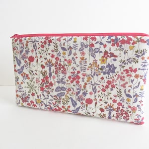 Può includere: Una pochette rettangolare con chiusura a zip su sfondo bianco, caratterizzata da una fitta stampa floreale nei toni del rosa, viola, giallo e verde. La pochette presenta una cerniera rosa brillante sul bordo superiore e una piccola linguetta.