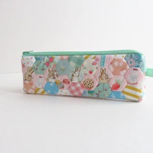 May include: A colorful pencil case with a hexagonal patchwork design. The case features a mint green zipper and pull tab. The fabric is patterned with bunnies, strawberries, milk bottles, and other whimsical illustrations.