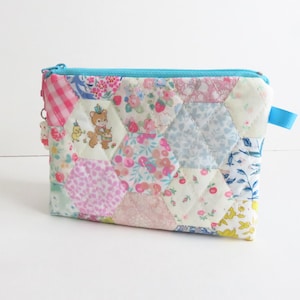 Puede incluir: Una bolsa con cremallera acolchada de patchwork con una cremallera azul y una cinta de cierre azul. La bolsa está hecha de varias telas florales y estampadas en tonos rosa, azul y blanco.