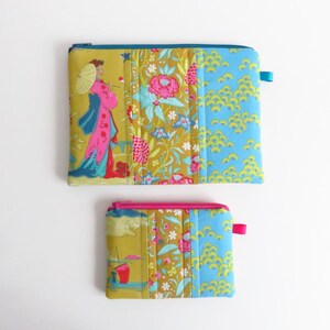 Può includere: Due pochette in tessuto trapuntato con chiusure a zip. La pochette più grande presenta una donna con un ombrello, motivi floreali e disegni di onde blu. La pochette più piccola ha motivi simili. Entrambe hanno accenti colorati.