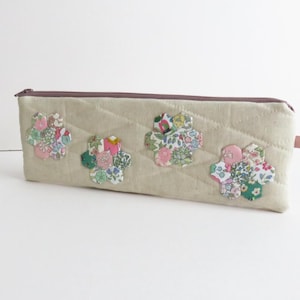 May include: A beige quilted pencil case with a brown zip and a small pink fabric loop. The case is decorated with four colourful floral appliqués. The flowers have pink, green, and blue patterns.