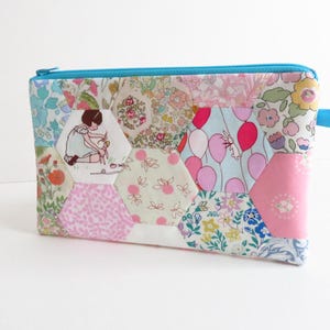 May include: A patchwork zippered pouch with a variety of floral and patterned fabrics in pink, blue, and white. The pouch features a light blue zipper and a small blue fabric loop. A hexagonal patch shows a girl with scissors.