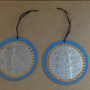 May include: Two round ornaments with blue borders and gold-colored embellishments. Each ornament has a silver center with handwritten symbols and a black ribbon for hanging. The ornaments are displayed on a light brown surface.