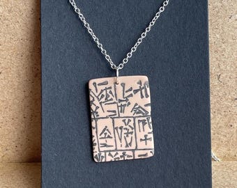 May include: A silver chain necklace with a rectangular pendant. The pendant is made of light pink ceramic with black markings that resemble ancient writing.