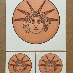 May include: A set of art prints featuring a stylized illustration of a face within a circle, surrounded by radiating lines. The face is in shades of orange and brown, with a detailed hairstyle and a serious expression. The set includes one large print and two smaller square prints.