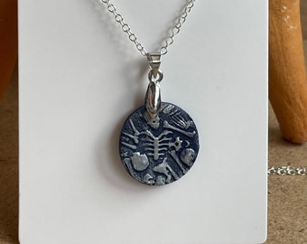 May include: A silver chain necklace with a round pendant featuring a silver skeleton and bones on a dark blue background.