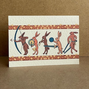 Può includere: Biglietto di auguri con un'illustrazione stravagante di conigli in abiti medievali. I conigli sono raffigurati con arco e frecce, un tamburo, una tromba, una spada e uno scudo. Il biglietto ha uno sfondo color crema con un bordo decorativo.