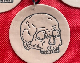 May include: A beige ceramic ornament with a black line drawing of a skull on it. The ornament is hanging from a black ribbon.