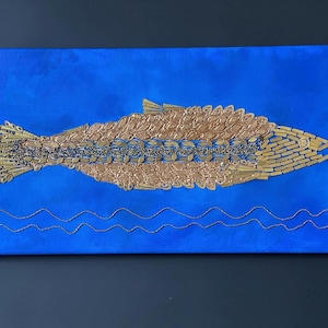 May include: A rectangular canvas art piece featuring a stylized fish design. The fish is composed of gold-colored embellishments against a vibrant blue background. Silver wavy lines are at the bottom, suggesting water.