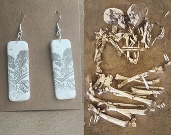 May include: A pair of white ceramic earrings with a gray print of two skeletons embracing. The earrings are rectangular and have silver hooks.