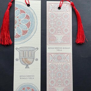May include: Two rectangular bookmarks with red tassels. One bookmark features a mosaic design in red, blue, and beige, with the text "KINGS WESTON ROMAN VILLA." The other bookmark has a geometric pattern in red and blue, also with the same text.