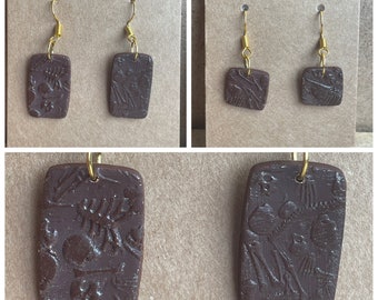 May include: A pair of brown clay earrings with a textured design. The earrings are rectangular in shape and have a gold hook.