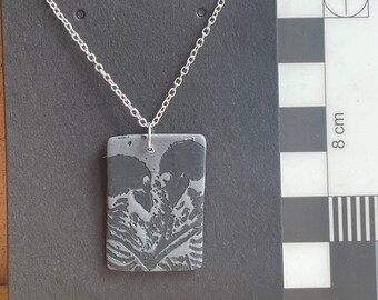 May include: A silver chain necklace with a black rectangular pendant featuring a gray and white abstract design.