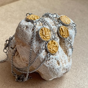 May include: A silver necklace with six circular, textured, gold-coloured pendants. The pendants are linked by fine chains and rest on a pale stone. The necklace has a clasp fastening.
