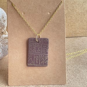 May include: A rectangular pendant necklace with a gold-colored chain. The pendant is a muted purple color and features an engraved design. The necklace is displayed on a brown card.