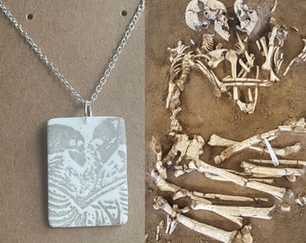 May include: A silver chain necklace with a rectangular pendant featuring a gray image of two skeletons embracing. The image is on a white background. To the right is a photo of two skeletons in the ground.