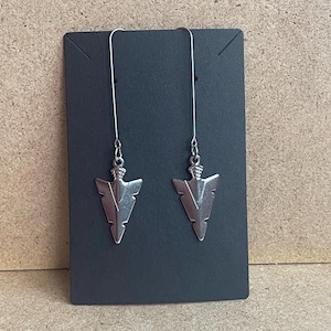 May include: A pair of silver arrowhead earrings with long, thin silver chains. The earrings are displayed on a black card.