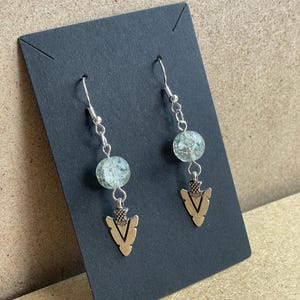 May include: A pair of silver-toned earrings with a beaded design and arrowhead pendants. The earrings feature a silver hook, a chain, a light green bead, and a silver arrowhead pendant. The earrings are displayed on a black card.
