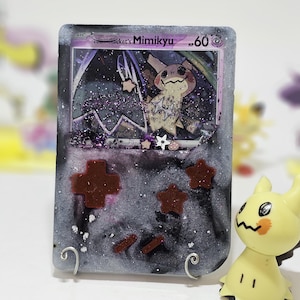 May include: A resin-coated Pokemon card featuring Mimikyu, with a gray and black background, red accents, and a yellow Mimikyu figure. The card has the text "Team Rocket's Mimikyu" and "HP 60".