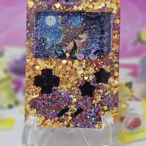 May include: A glittery, decorative Game Boy-shaped resin piece. The frame is gold with a mix of glitter colors. The screen shows a night scene with a witch and a full moon. The buttons are black.