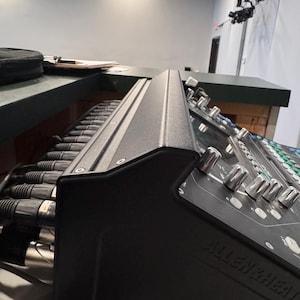 May include: A black Allen & Heath audio mixer with silver knobs and a row of black cables connected to the back.