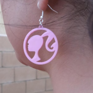 Può includere: Orecchini circolari rosa con la silhouette del profilo di una donna con la coda di cavallo. Gli orecchini sono appesi a un gancio d'argento. La persona indossa una maglietta rosa. Lo sfondo è un muro di mattoni.