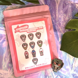 May include: A pink packet containing a card with several enamel pins. The card reads "Charriiiiel Seeker Enamel Pin Blind Bag" and "Collect Them All!". One pin is outside the packet, showing a robot design in grey, red, and white.