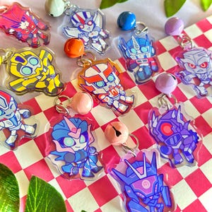May include: A collection of acrylic charms featuring various stylized robot characters in vibrant colors, each attached to a small metal jump ring and a colorful jingle bell. The charms display diverse designs including yellow, blue, red, and purple figures.