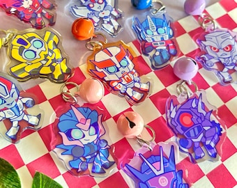 Könnte beinhalten: Eine Sammlung von Acryl-Anhängern mit verschiedenen stilisierten Roboterfiguren in leuchtenden Farben, die jeweils an einem kleinen Metallring und einer bunten Glocke befestigt sind. Die Anhänger zeigen diverse Designs in Gelb, Blau, Rot und Violett.