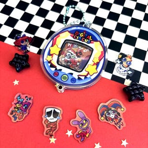 May include: A round Digital Circus themed keychain with a screen displaying a character, accompanied by several small acrylic character charms and black star-shaped beads. The keychain features a blue border with yellow stars and a metal chain.