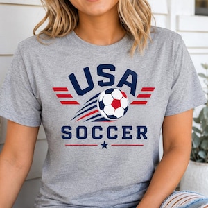 May include: A grey t-shirt with the text "USA SOCCER" in navy blue and red, featuring a soccer ball graphic. The shirt has a crew neck and short sleeves. The design includes a star and wing-like elements.