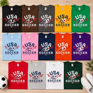 May include: A selection of USA Soccer t-shirts in diverse colours, such as black, brown, grey, yellow, and green. Each shirt displays the text "USA SOCCER" with a soccer ball graphic and star accents. The shirts are laid out flat.