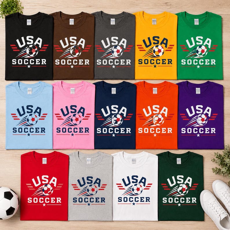 May include: A selection of USA Soccer t-shirts in diverse colours, such as black, brown, grey, yellow, and green. Each shirt displays the text "USA SOCCER" with a soccer ball graphic and star accents. The shirts are laid out flat.