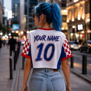 Puede incluir: Una camiseta corta blanca de manga corta con un patr&oacute;n de cuadros rojos y blancos en los laterales y detalles azules. La parte trasera muestra el texto YOUR NAME en azul sobre el n&uacute;mero 10 grande en azul. La prenda se combina con vaqueros azul claro.