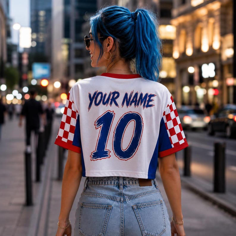 Puede incluir: Una camiseta corta blanca de manga corta con un patr&oacute;n de cuadros rojos y blancos en los laterales y detalles azules. La parte trasera muestra el texto YOUR NAME en azul sobre el n&uacute;mero 10 grande en azul. La prenda se combina con vaqueros azul claro.