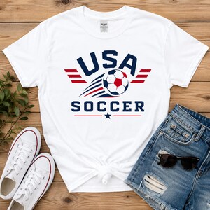 May include: White t-shirt with a USA Soccer graphic. The design features the text "USA SOCCER" in navy blue, a soccer ball, and red and blue accents. The shirt is paired with denim shorts and white trainers.