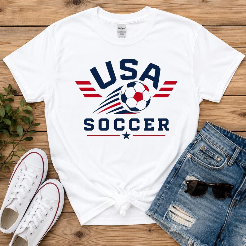 May include: White t-shirt with a USA Soccer graphic. The design features the text "USA SOCCER" in navy blue, a soccer ball, and red and blue accents. The shirt is paired with denim shorts and white trainers.