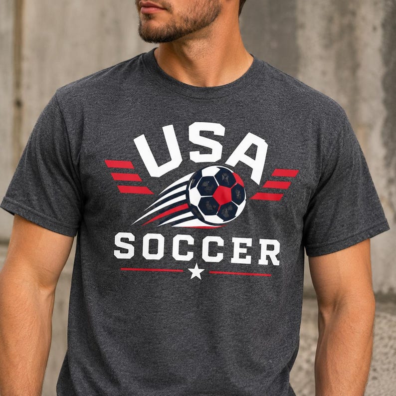 May include: Dark grey t-shirt featuring the text "USA SOCCER" in white, red, and black. The design includes a soccer ball with red and black panels and white streaks, and red wing-like accents. A star is below the word "SOCCER".